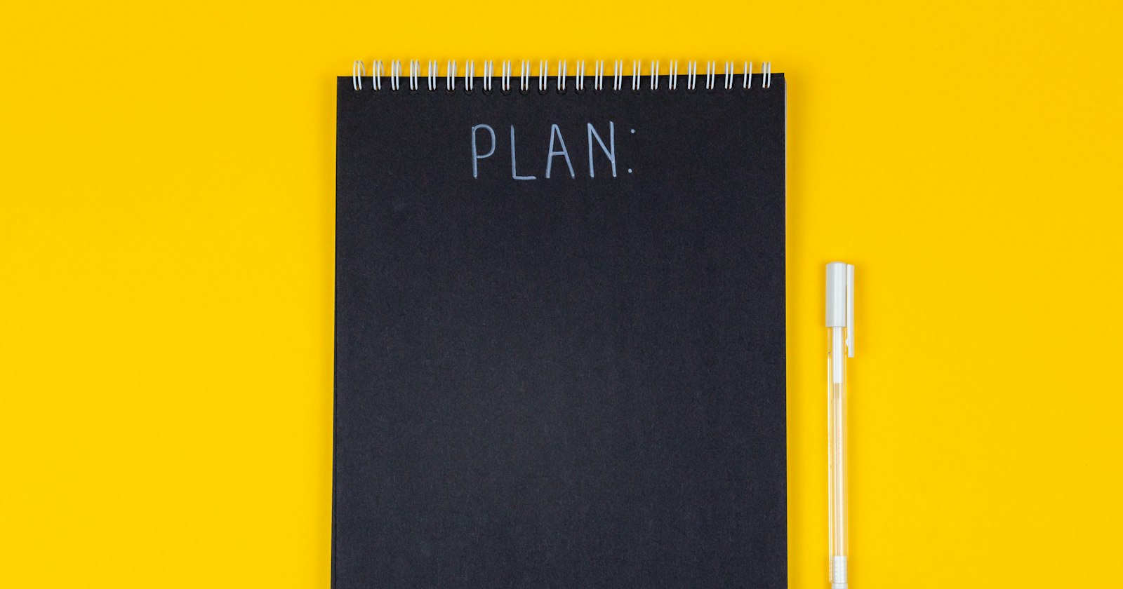 Caderno preto com a palavra PLAN sobre fundo amarelo, ao lado de uma caneta