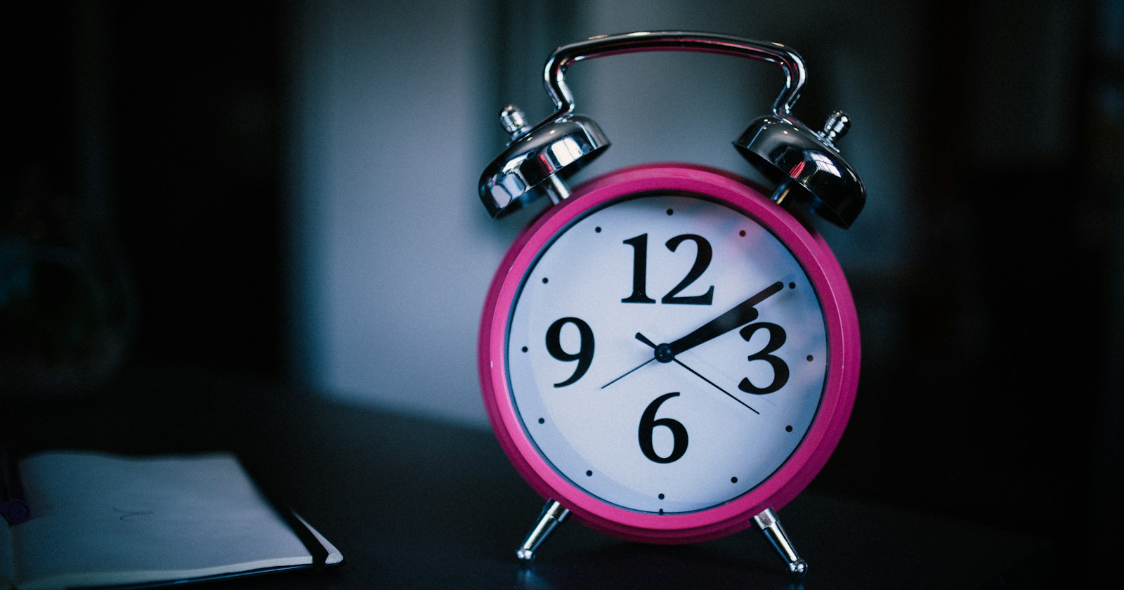 Despertador rosa sobre uma mesa ao lado de um caderno